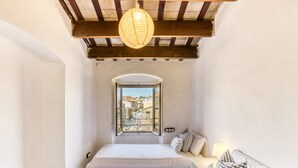 A bedroom with a bed, a bedside table with a lamp, a window with a view of buildings, and a hanging light fixture. - Modernist Manor House for 14 – 7 Bedrooms – Central Palafrugell – Patio – Garage (Palafrugell)