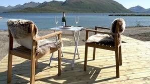 A wooden deck with two fur-covered chairs facing a table set with a bottle and glasses, overlooking a calm lake and distant mountains. - Arctic Glamping-Dome (Andoy)