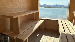 A wooden cabin with a bench and a table near a window overlooking a body of water and mountains. - Arctic Glamping-Dome (Andoy)