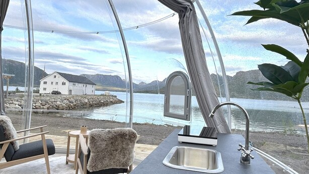 A modern kitchen with a large window overlooking a lake and a white building. - Arctic Glamping-Dome (Andoy)