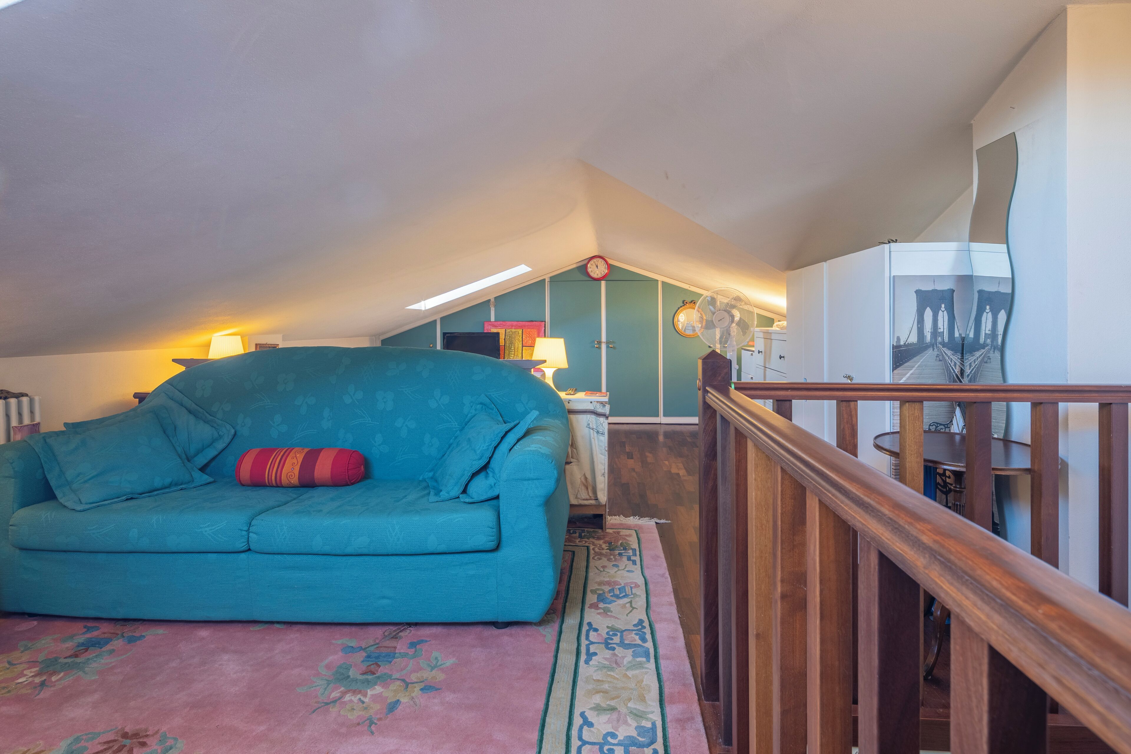 A teal sofa with a red pillow, a wooden staircase, and a wall with a clock and a fan.