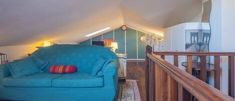 A teal sofa with a red pillow, a wooden staircase, and a wall with a clock and a fan.