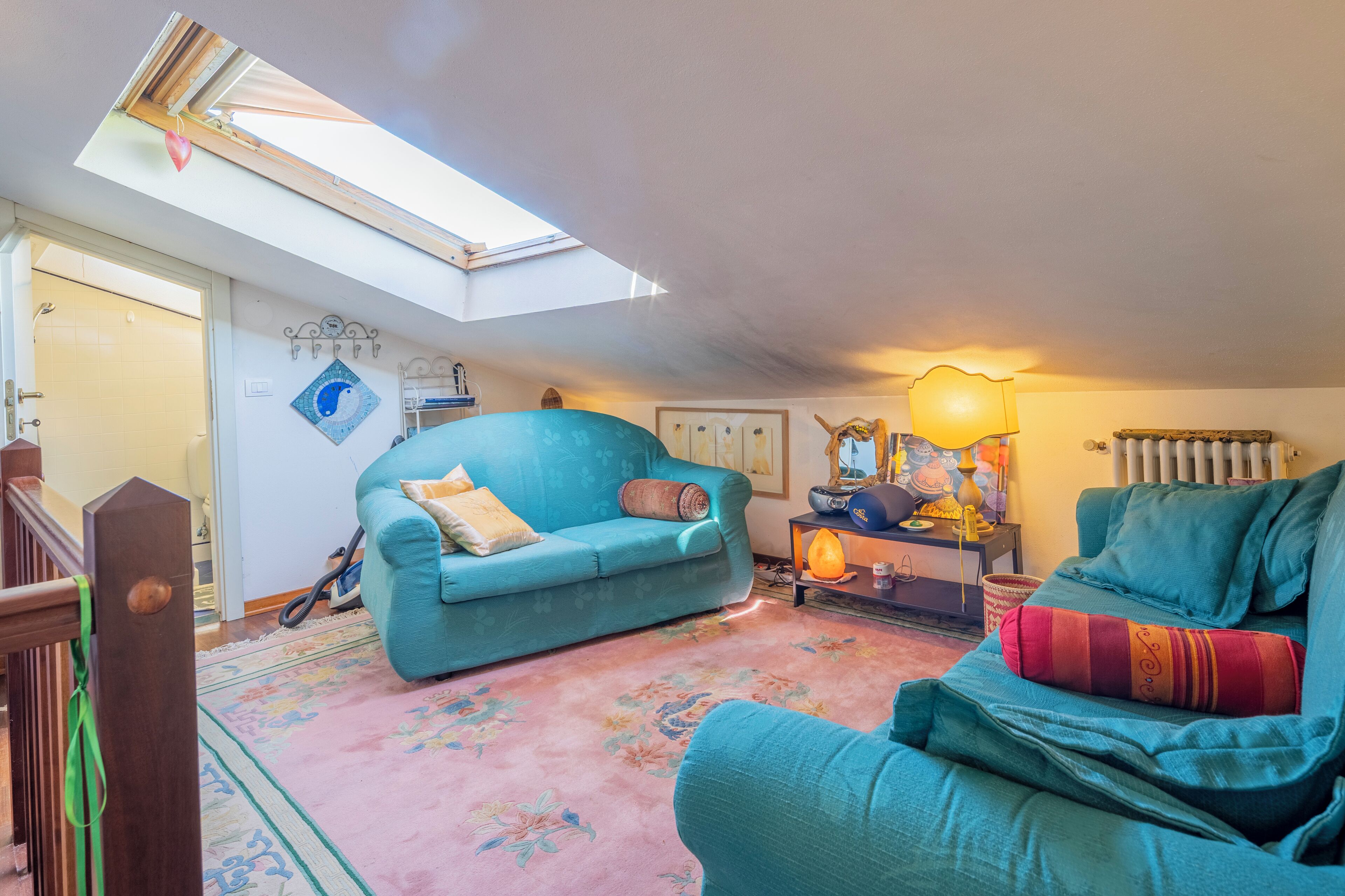A cozy living room with a teal sofa, a pink floral rug, and a skylight above.
