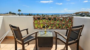Une terrasse avec deux fauteuils en osier et une table, d’où l’on peut admirer un paysage côtier avec des bâtiments et des palmiers.
