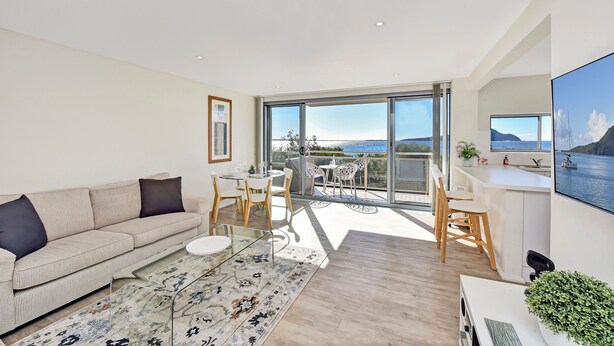 Ein modernes Wohnzimmer mit einer Couch, einem Couchtisch und einem Essbereich mit Stühlen und einem Tisch. Ein großes Fenster bietet Blick aufs Meer.