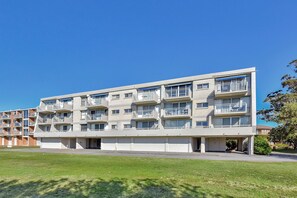 A multi-story residential building with balconies, a flat roof, and a grassy area in front.
