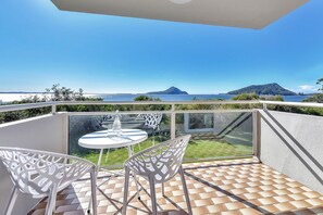 A balcony with a glass table and chairs, overlooking a body of water and a distant mountain.