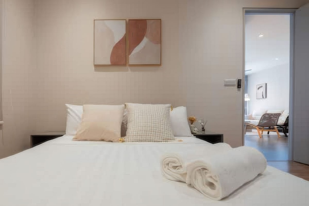 Un lit bien fait, avec du linge de lit blanc et deux oreillers, une petite table de chevet avec une lampe, et une œuvre d’art abstraite accrochée au mur.