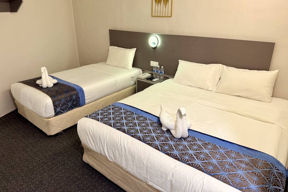 Een hotelkamer met twee bedden, elk met een witte deken, een blauw gebedrukte sprei en een witte handdoek. Er staat een nachtkastje met een lamp, een telefoon en twee waterflessen.