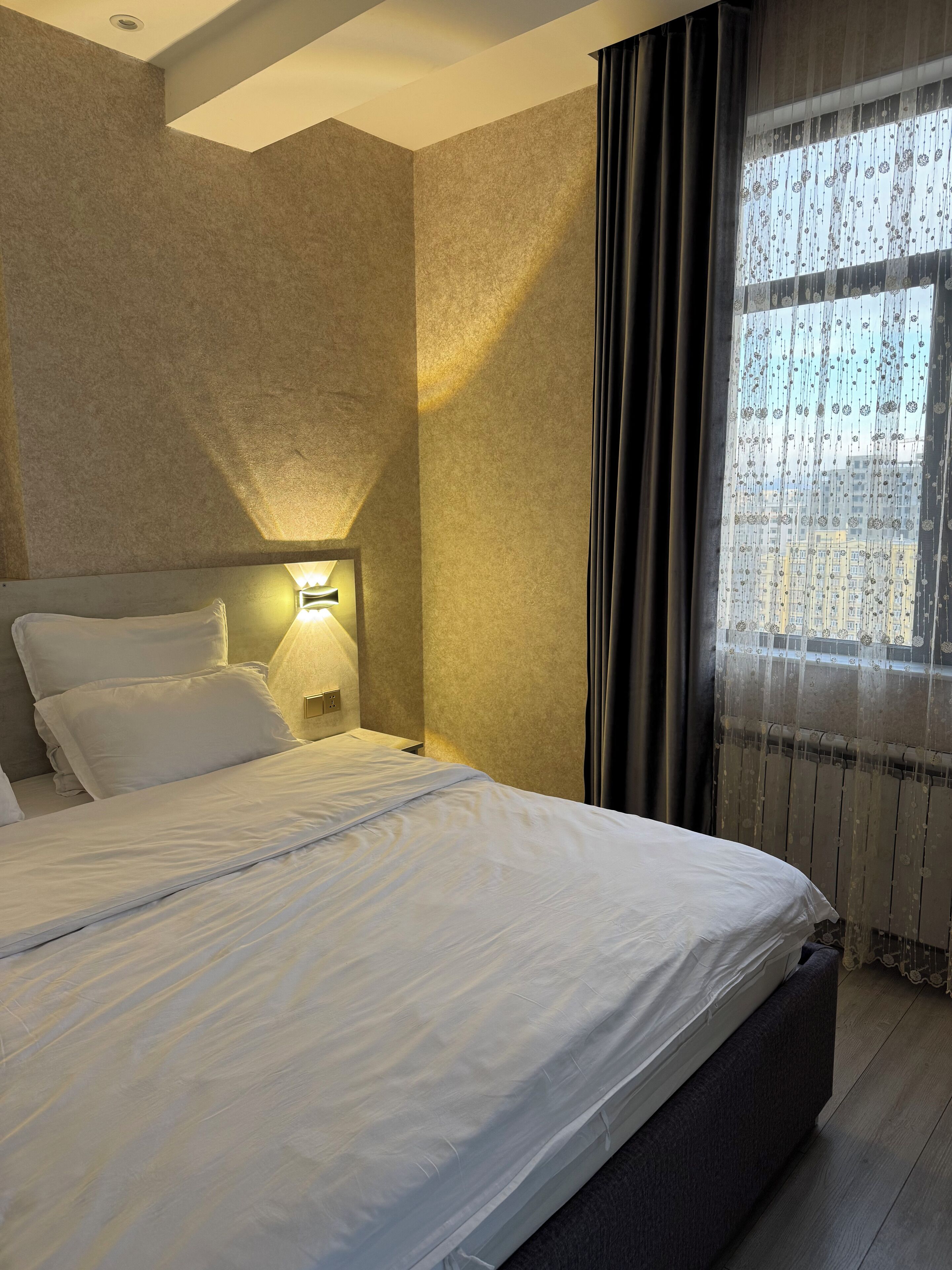 Um quarto com cama, mesa de cabeceira, luminária na parede e janela com cortinas.