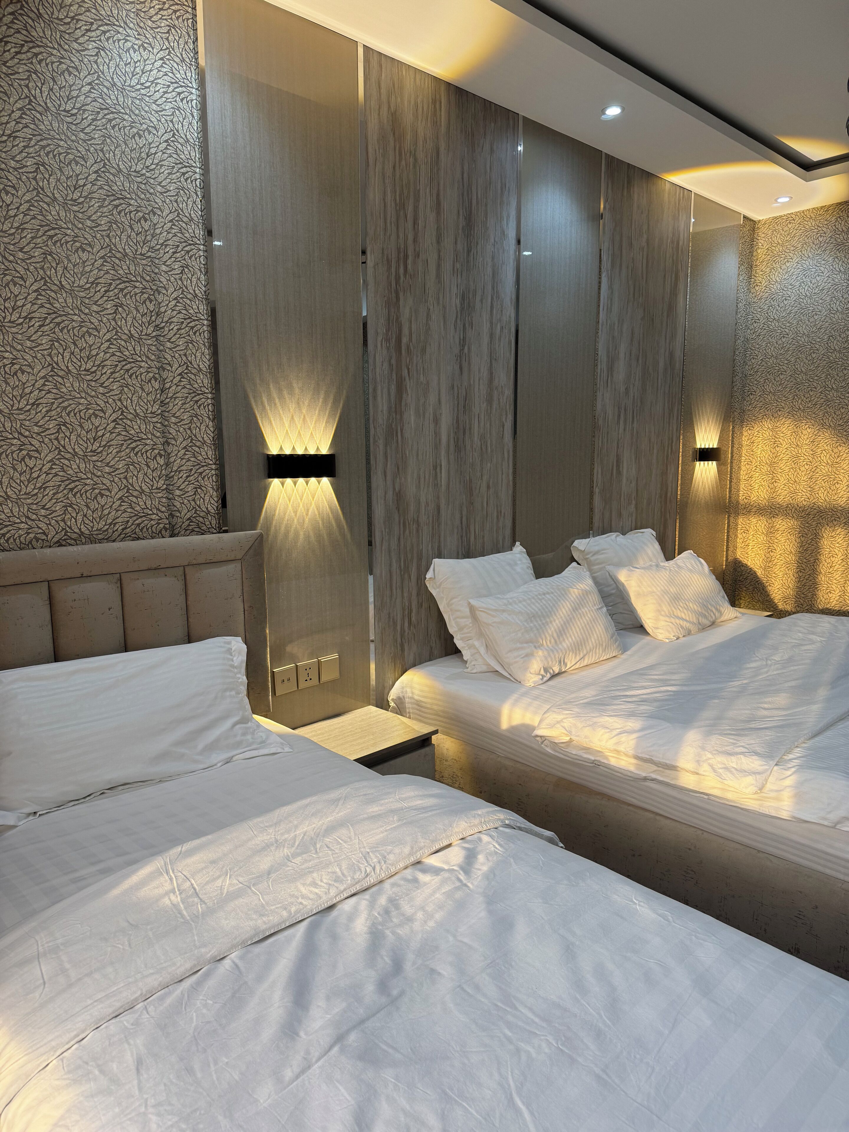 Een moderne slaapkamer met twee bedden, een houten hoofdbord en wandlampen.