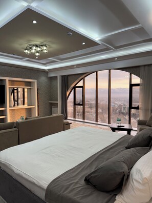 A modern hotel room with a large bed, a seating area, a vanity with a mirror, and a view of a cityscape through a large window. - Panorama Hotel (Dushanbe)