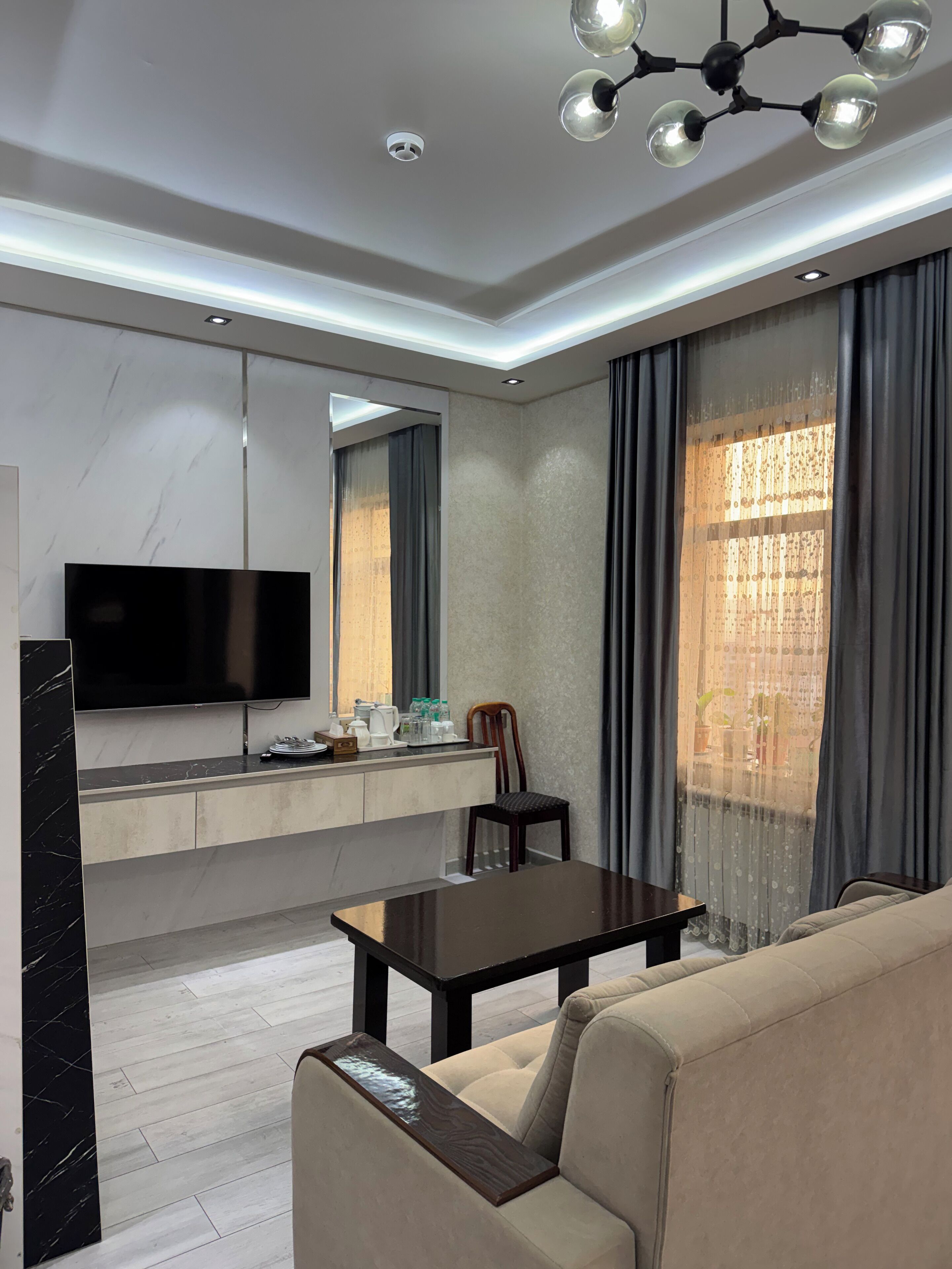 A modern living room with a flat-screen TV, a dining table, a sofa, and a chandelier.