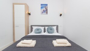 Cama bem arrumada com duas almofadas, um cobertor cinza e duas toalhas dobradas. Uma porta de madeira à esquerda e um quadro pendurado na parede acima da cama.