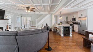 Un vaste salon doté d’un canapé d’angle gris, d’un ventilateur de plafond et d’un coin cuisine avec des meubles blancs et des appareils électroménagers en acier inoxydable.