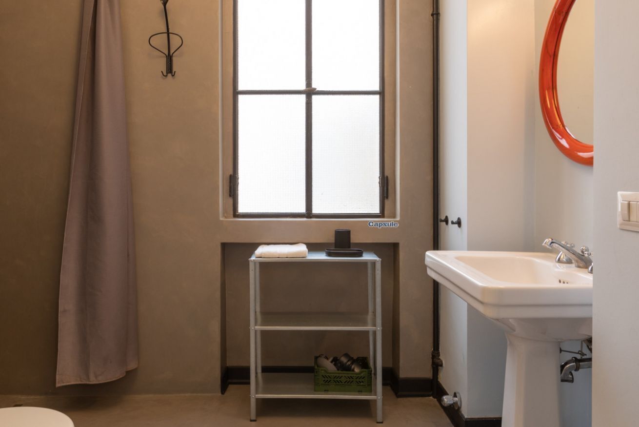 Ein Badezimmer mit Fenster, einem weißen Waschbecken, einem Metallregal mit Schuhen und einem Handtuch an der Wand.