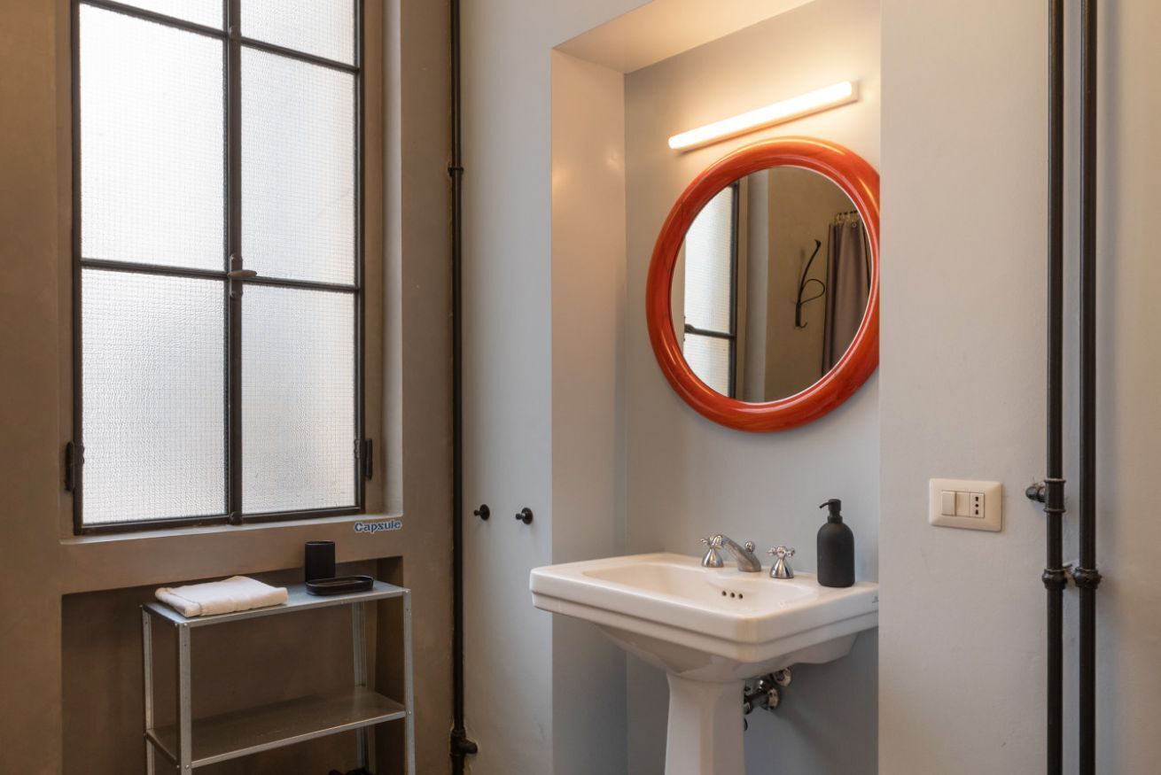 A bathroom with a white sink, a round mirror with an orange frame, a window with frosted glass, and a shelf with a towel and a black object.