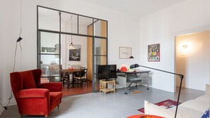 A modern living room with a red armchair, a TV on a wooden stand, a desk with a computer, and a dining area visible through a glass partition. - Splendid two-room apartment Dateo - Mirable PM (Milano)