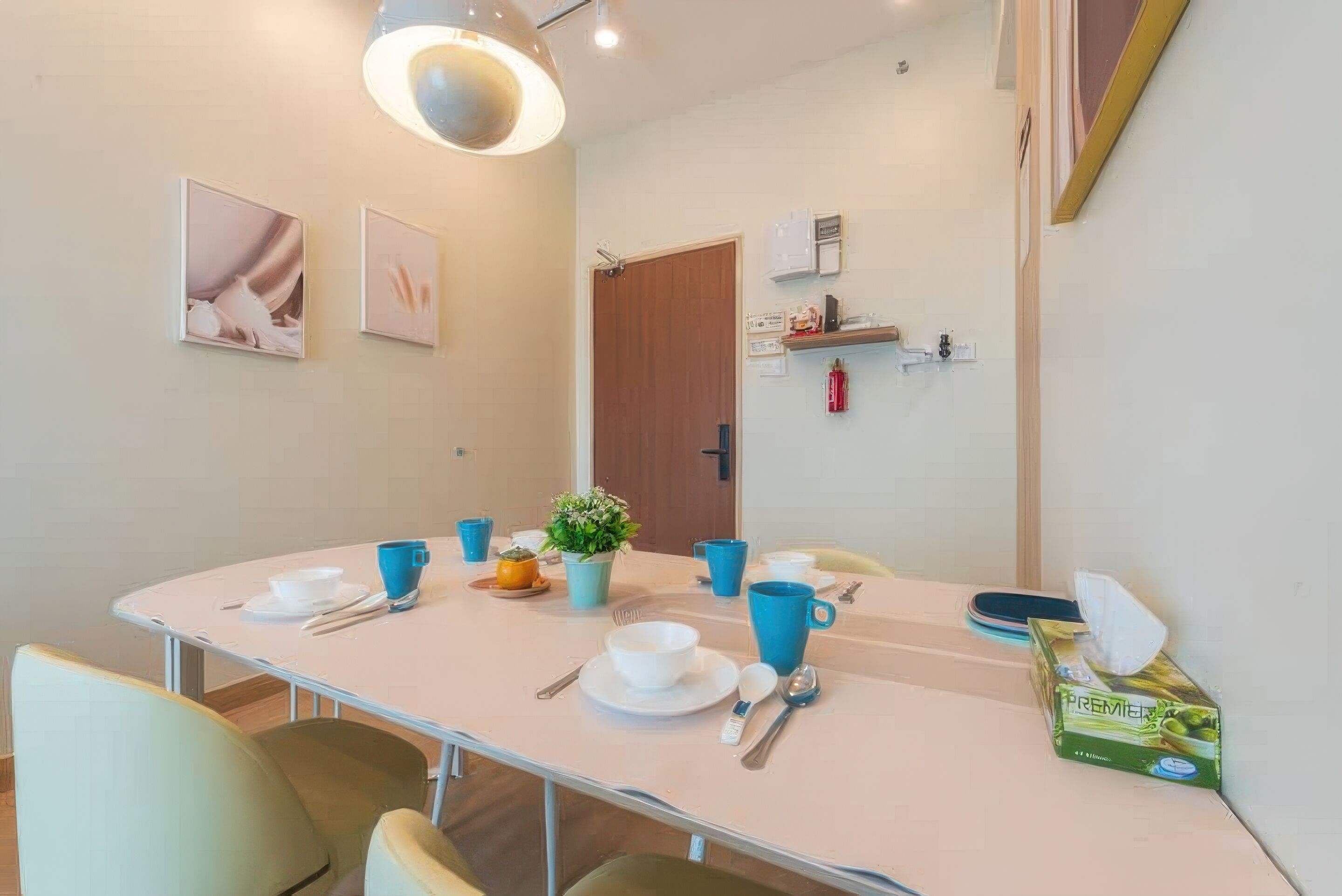 Comedor con una mesa dispuesta para cuatro, que incluye tazas azules, platos blancos y cubiertos. Hay cuadros en la pared, una planta pequeña en la mesa y un extintor montado en la pared.