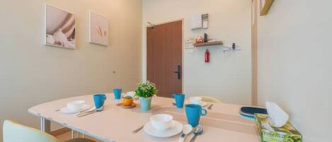 Comedor con una mesa dispuesta para cuatro, que incluye tazas azules, platos blancos y cubiertos. Hay cuadros en la pared, una planta pequeña en la mesa y un extintor montado en la pared.