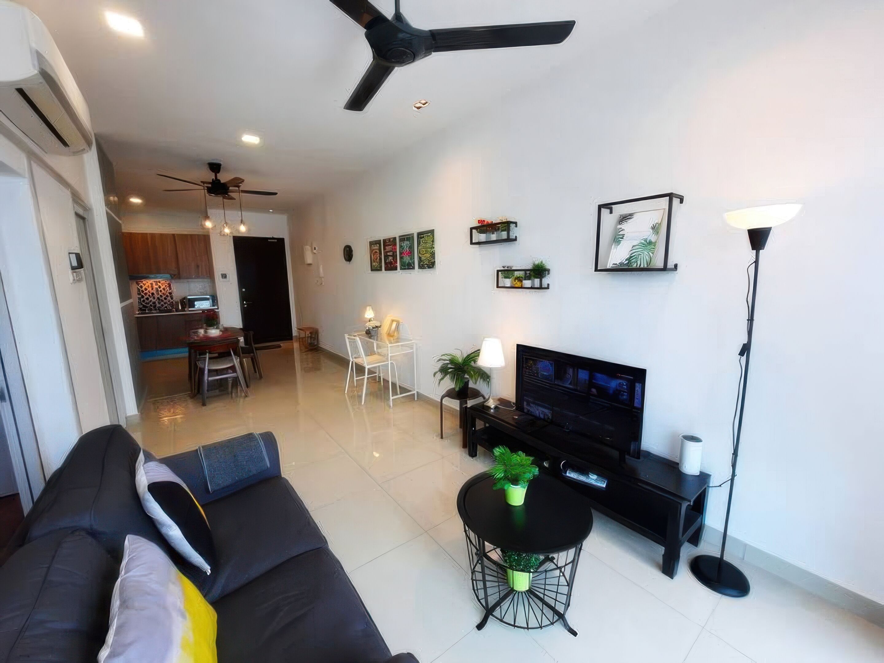 A living room with a black sofa, a TV, a small round table, and a ceiling fan. There is a kitchen in the background with wooden cabinets and a dining area.