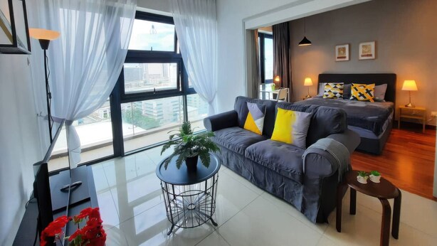 Une chambre d’hôtel moderne avec un canapé, une petite table agrémentée d’une plante, un lit avec des coussins à motifs et une vue sur la ville grâce à de grandes fenêtres.