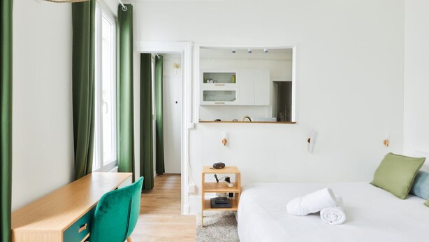 A modern hotel room with a bed, a desk, a chair, a mirror, and a small shelf.