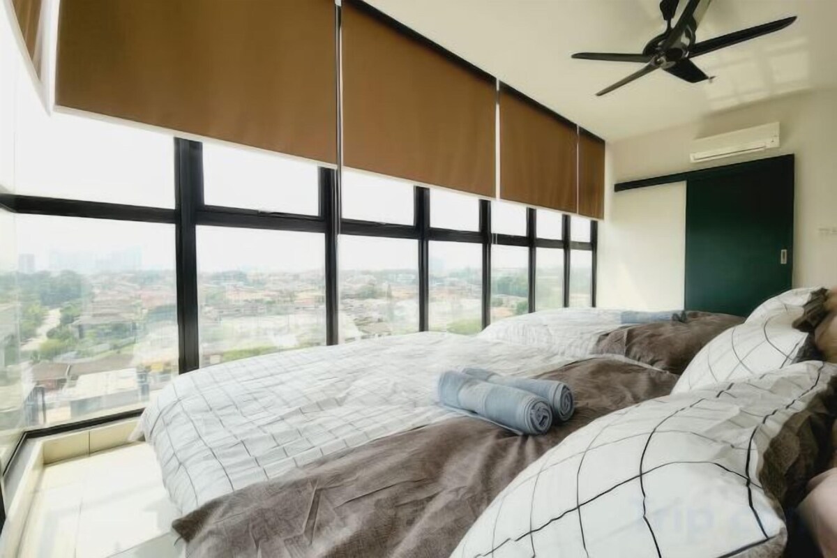 Une chambre avec un grand lit, un ventilateur de plafond et une vue sur le paysage urbain, visible à travers de grandes fenêtres.
