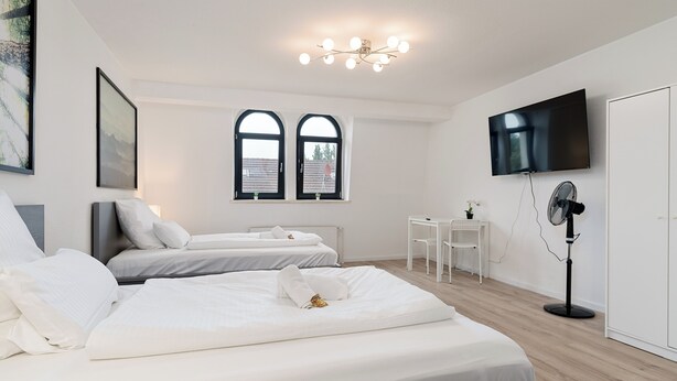 A modern hotel room with two beds, a desk, a chair, a TV, and a standing fan. - Big groups / Families / Exhibition (Duisburg)