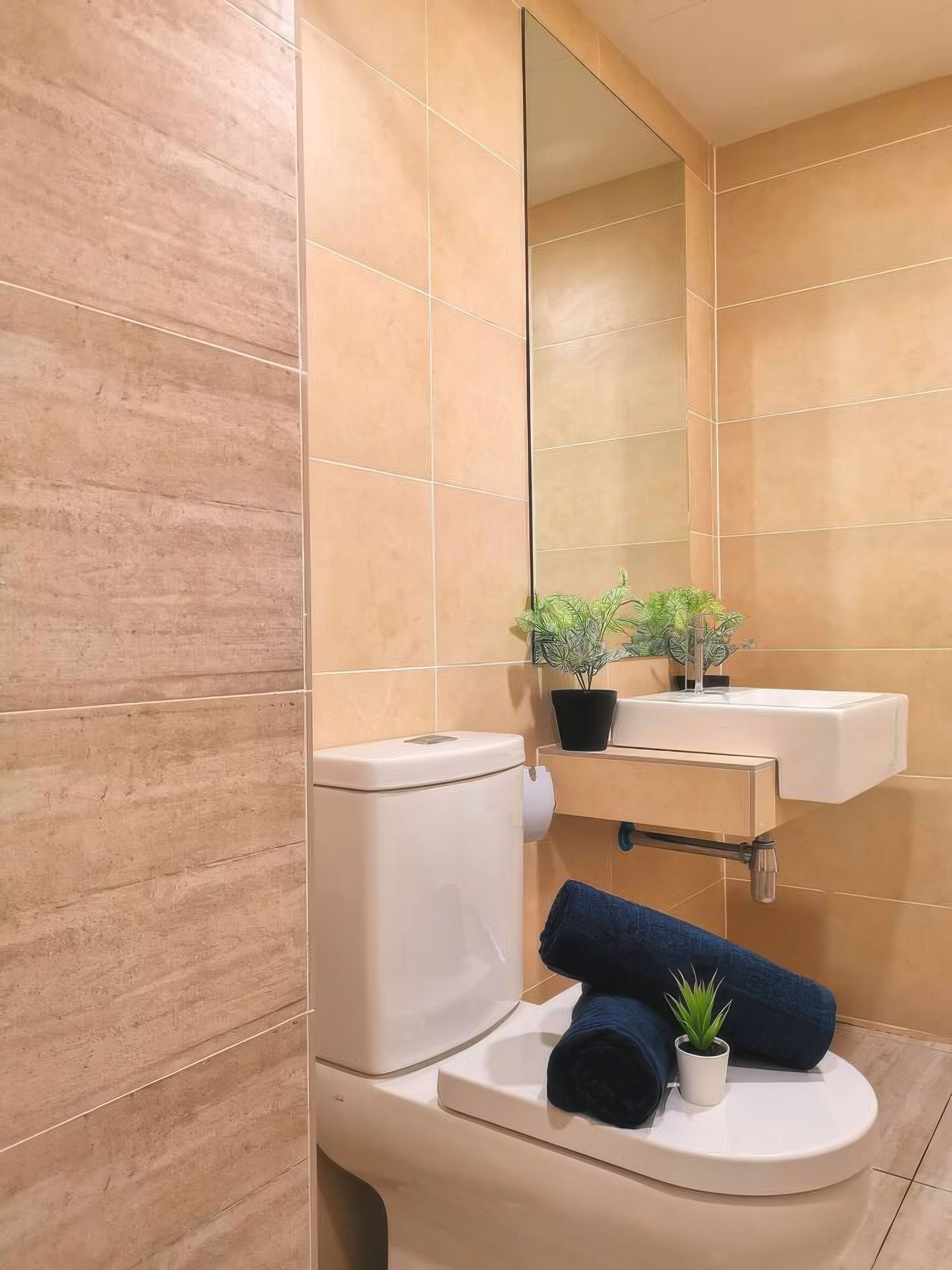 Un baño con baldosas color beige, un inodoro blanco, un lavamanos con un espejo encima y dos plantas en macetas.