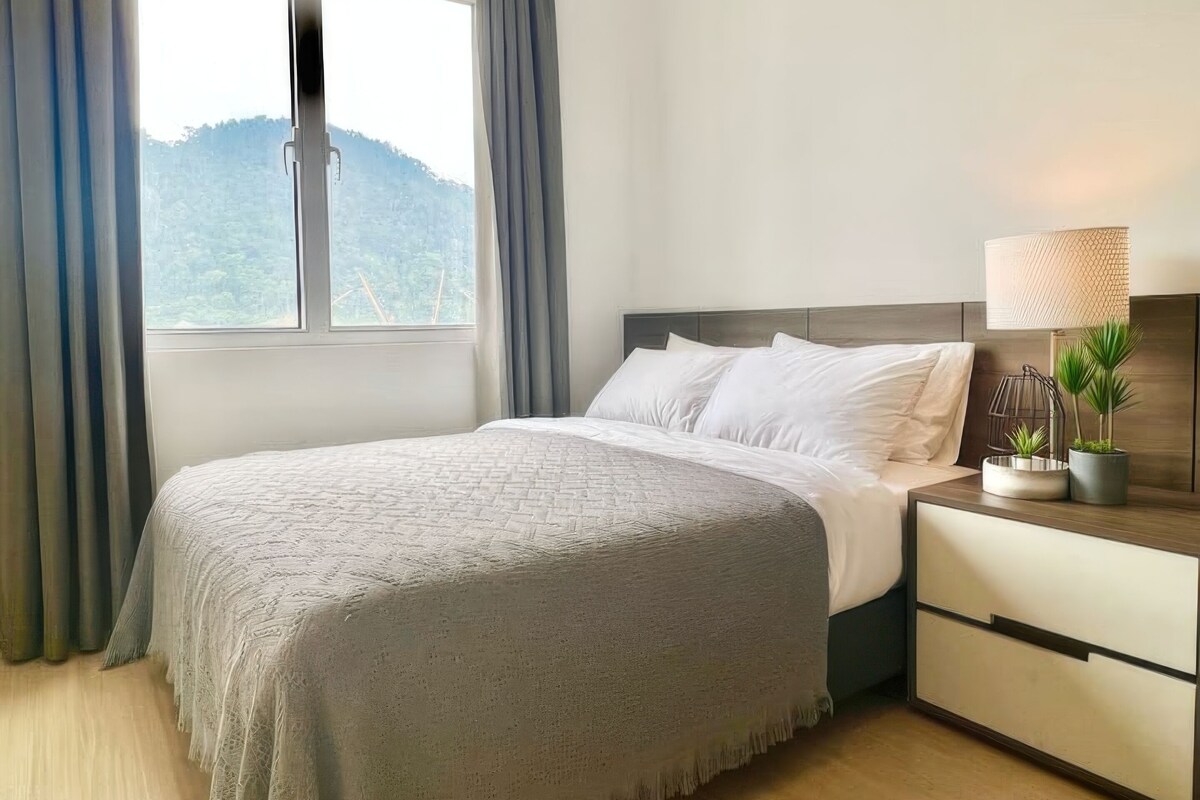 Un lit bien fait, recouvert d’une couverture grise, une table de chevet avec une lampe et une petite plante, et une fenêtre avec des rideaux.