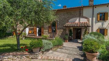 A stone building with a tiled roof, a covered outdoor seating area, and a garden with potted plants and a tree.
