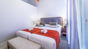 A hotel room with a bed, a bench, a nightstand, a lamp, and a window with curtains.