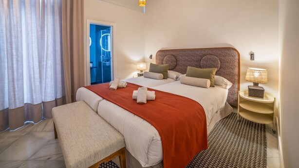 A hotel room with a large bed, a bench, a bedside table with a lamp, and a view of the outside.