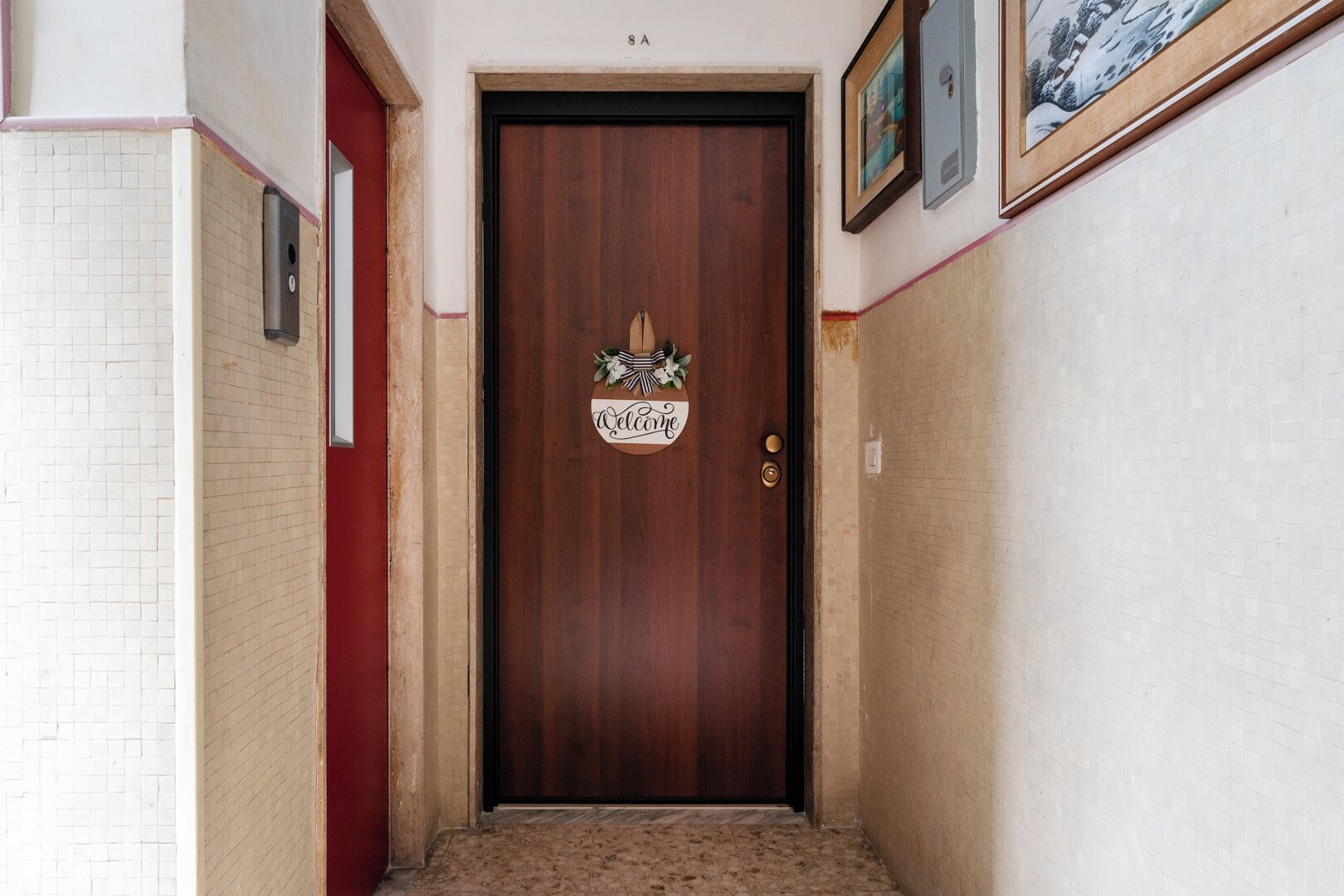 A narrow hallway with a wooden door at the end, a welcome sign, and a framed picture above the door.