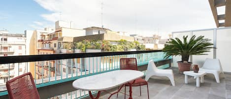Un balcón con mesa y sillas, plantas en macetas y vistas de edificios circundantes.