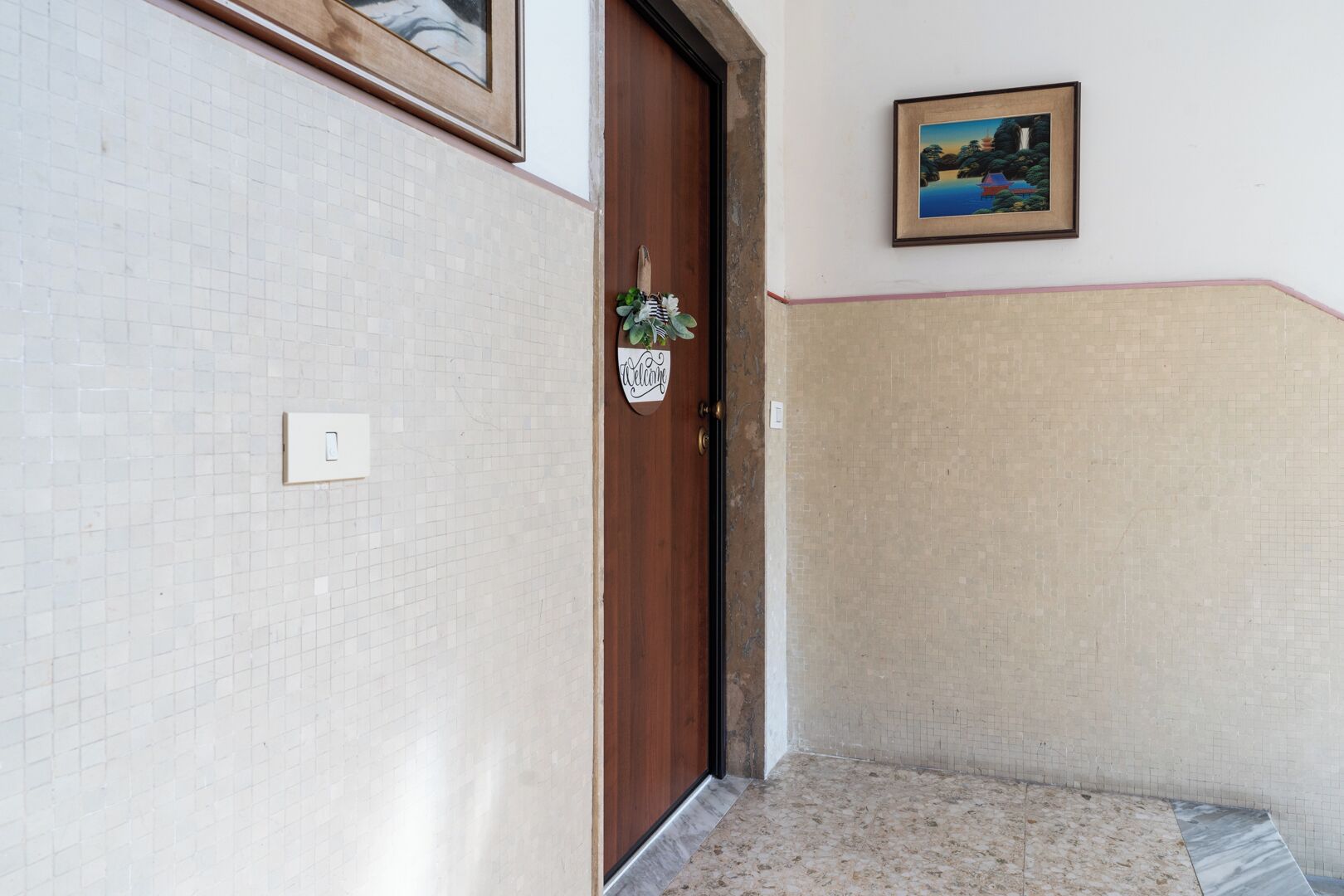 A hallway with a tiled wall, a wooden door, a welcome sign, and a framed picture on the wall.