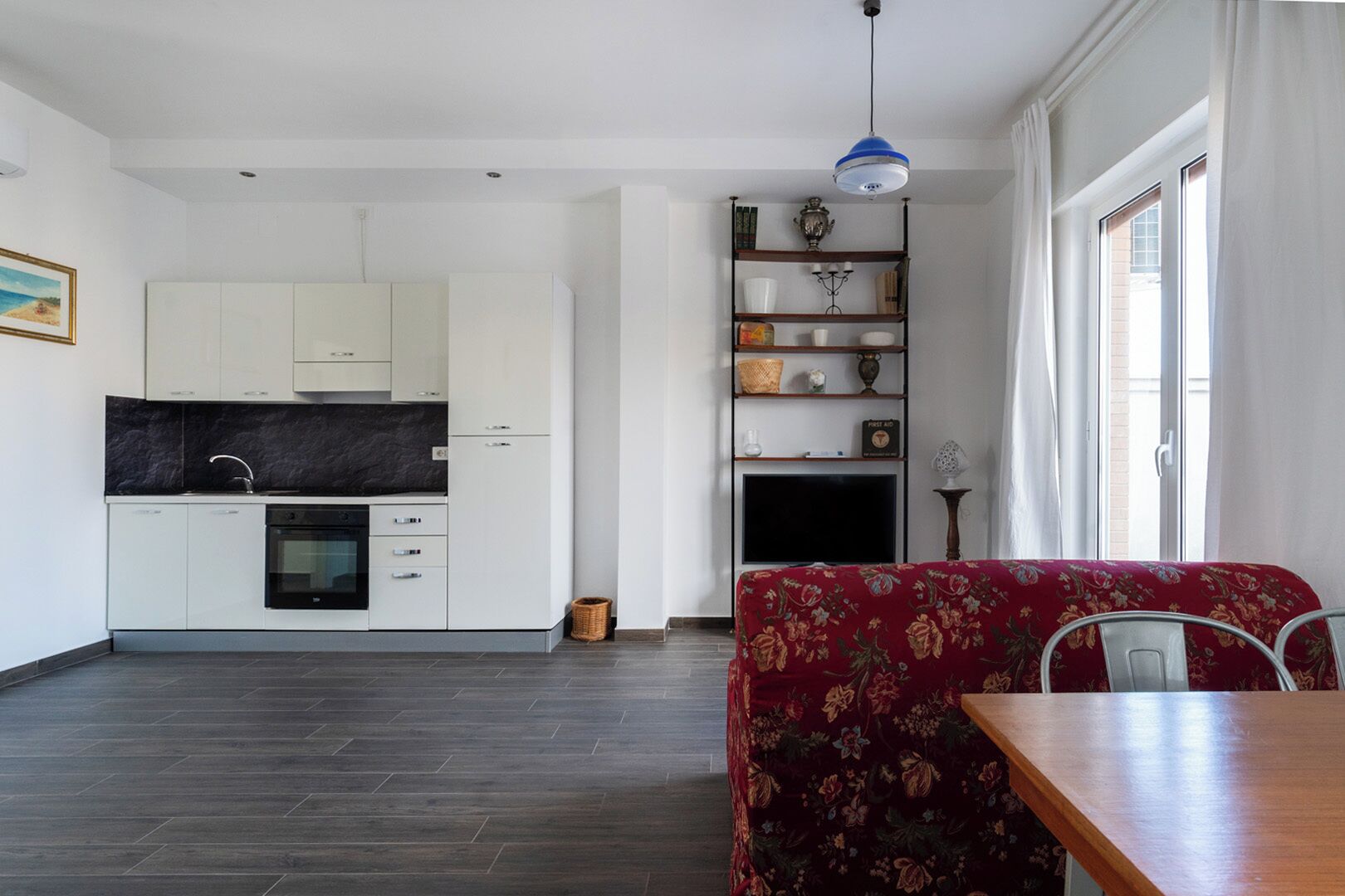 Een moderne keuken met witte kasten en een zwart aanrecht, een zithoek met een rode sofa met bloemenprint, en een televisie op een plank.