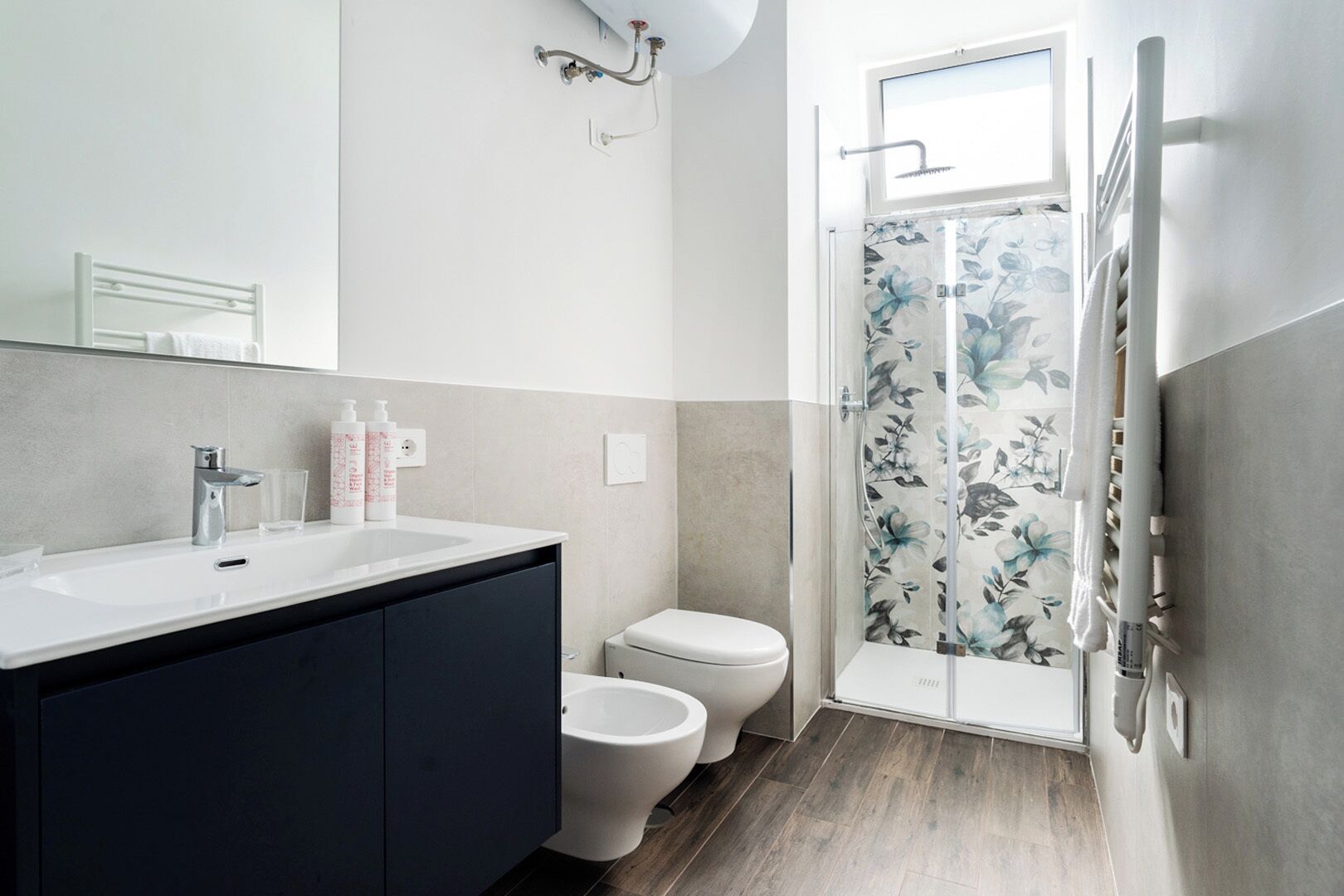 Een moderne badkamer met een wit wastafelmeubel, donkere kastjes, een toilet en een douche met bloemenprint gordijnen.