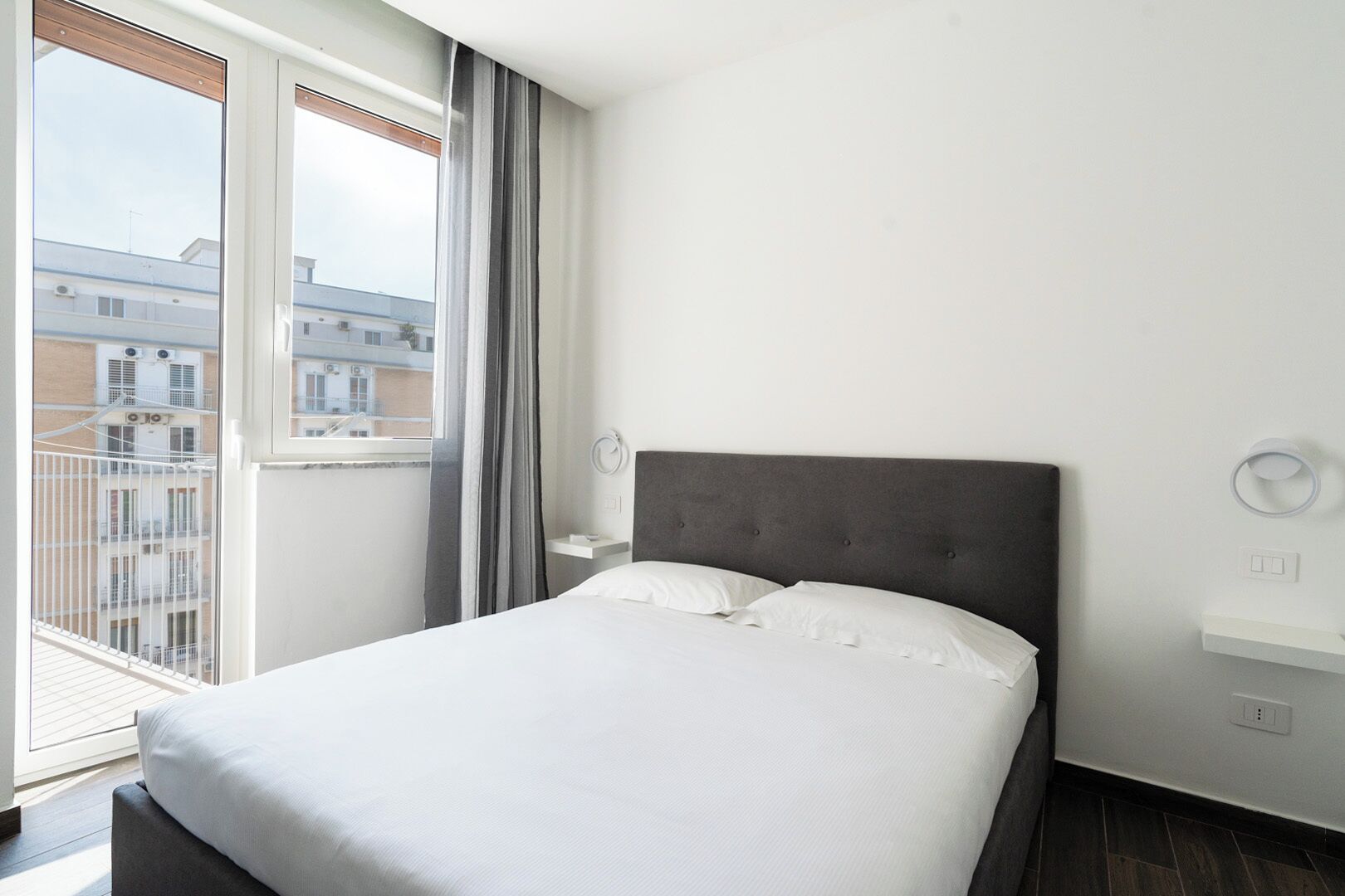 Une chambre d’hôtel avec un grand lit, une table de chevet, une lampe fixée au mur et une vue sur les bâtiments par la fenêtre.