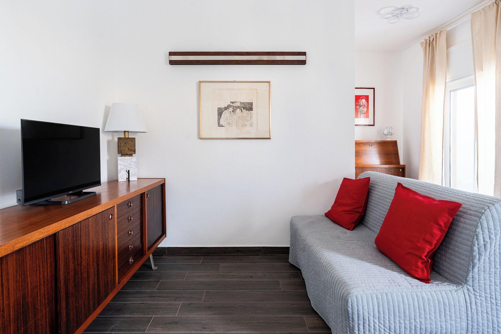 Ein modernes Wohnzimmer mit Flachbildfernseher, Holzschrank, grauem Sofa mit roten Kissen, gerahmtem Kunstwerk und Couchtisch aus Holz.