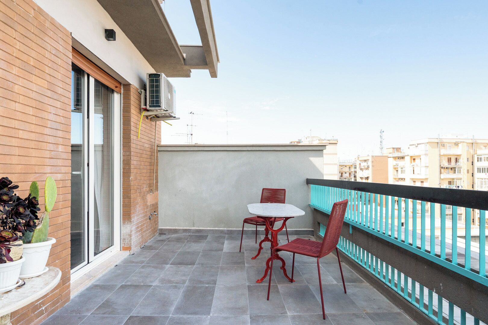 Un balcón con una mesa, sillas, una planta en maceta y vistas de edificios al fondo.