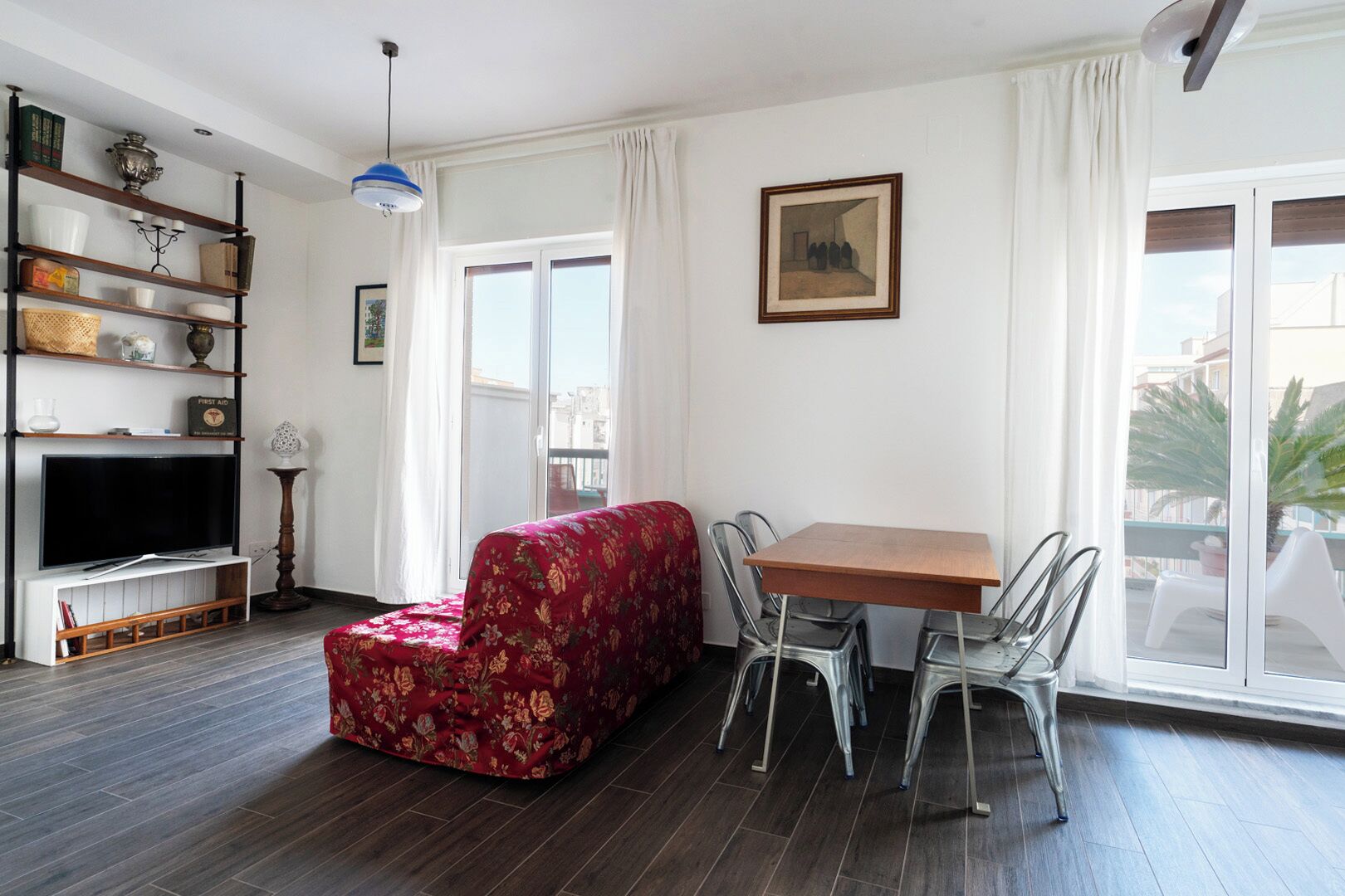 A living room with a TV, a red floral sofa, a dining table with chairs, and a large window with curtains.