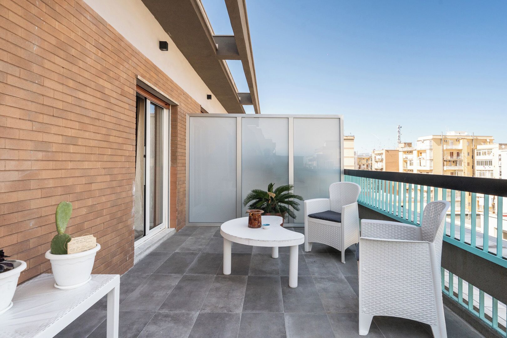 Uma varanda com mesa branca e cadeiras de vime, um cacto em vaso, e vista para edifícios.