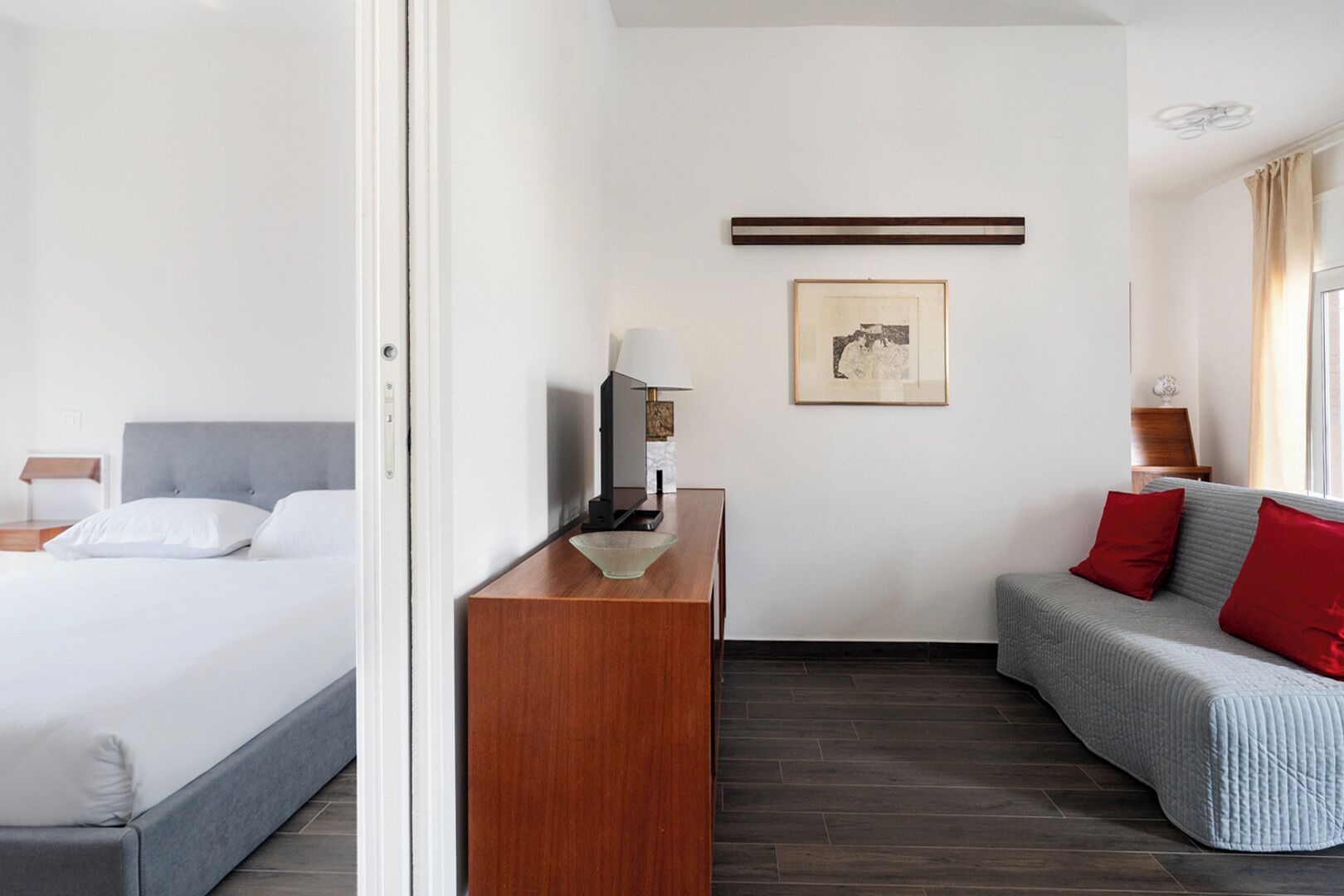 A hotel room with a bed, a wooden side table with a lamp, a grey sofa with red cushions, and a framed picture on the wall.