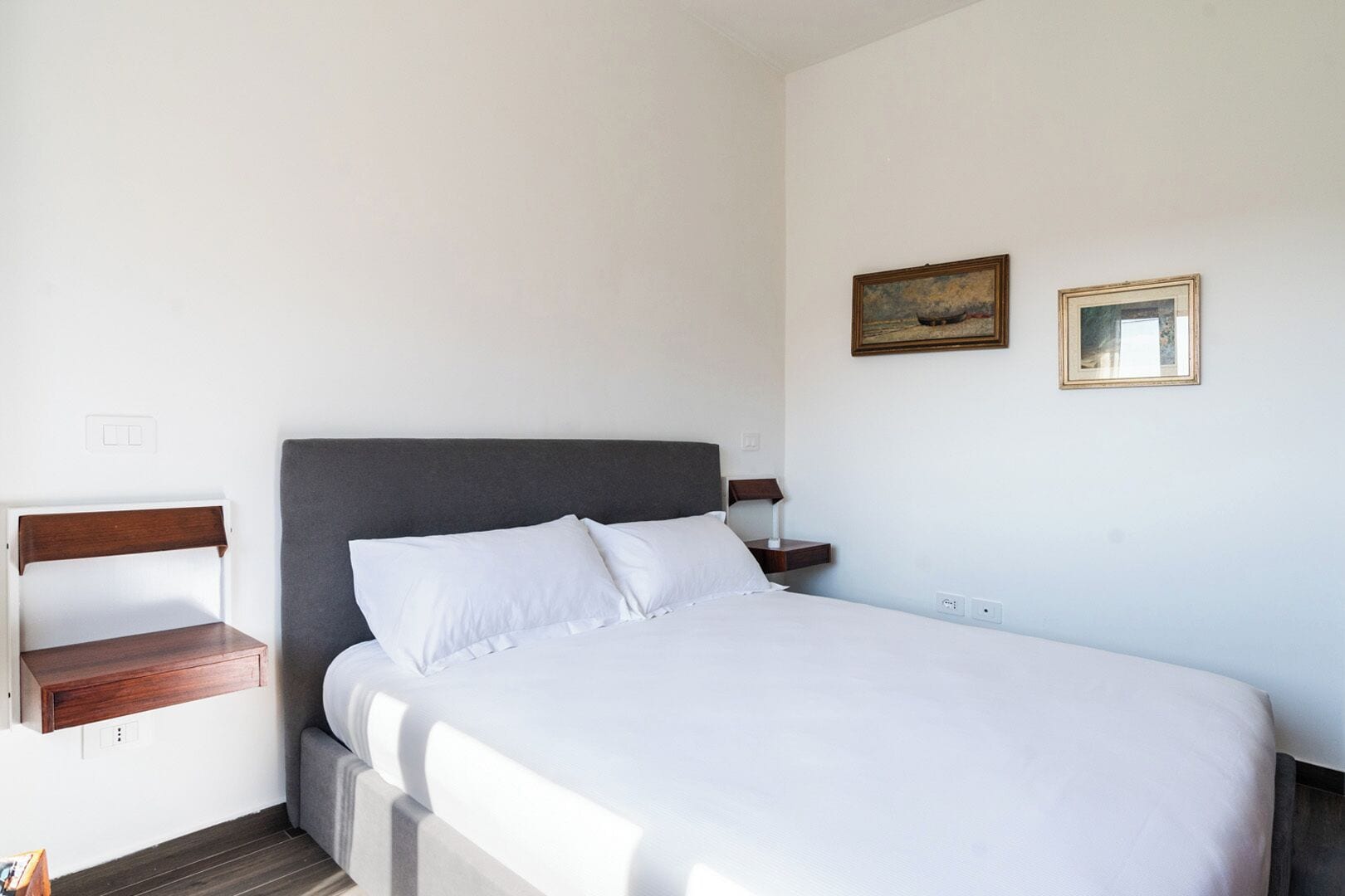 Una cama bien hecha con ropa blanca, un cabecero oscuro y una mesita de noche con una lámpara. Hay dos cuadros enmarcados en la pared.