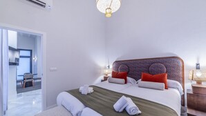 Ein modernes Hotelzimmer mit einem großen Bett, einer grünen Tagesdecke und ordentlich gefalteten Handtüchern. In der Ecke steht ein kleiner Tisch mit Stuhl, und auf dem Nachttisch steht eine Lampe.