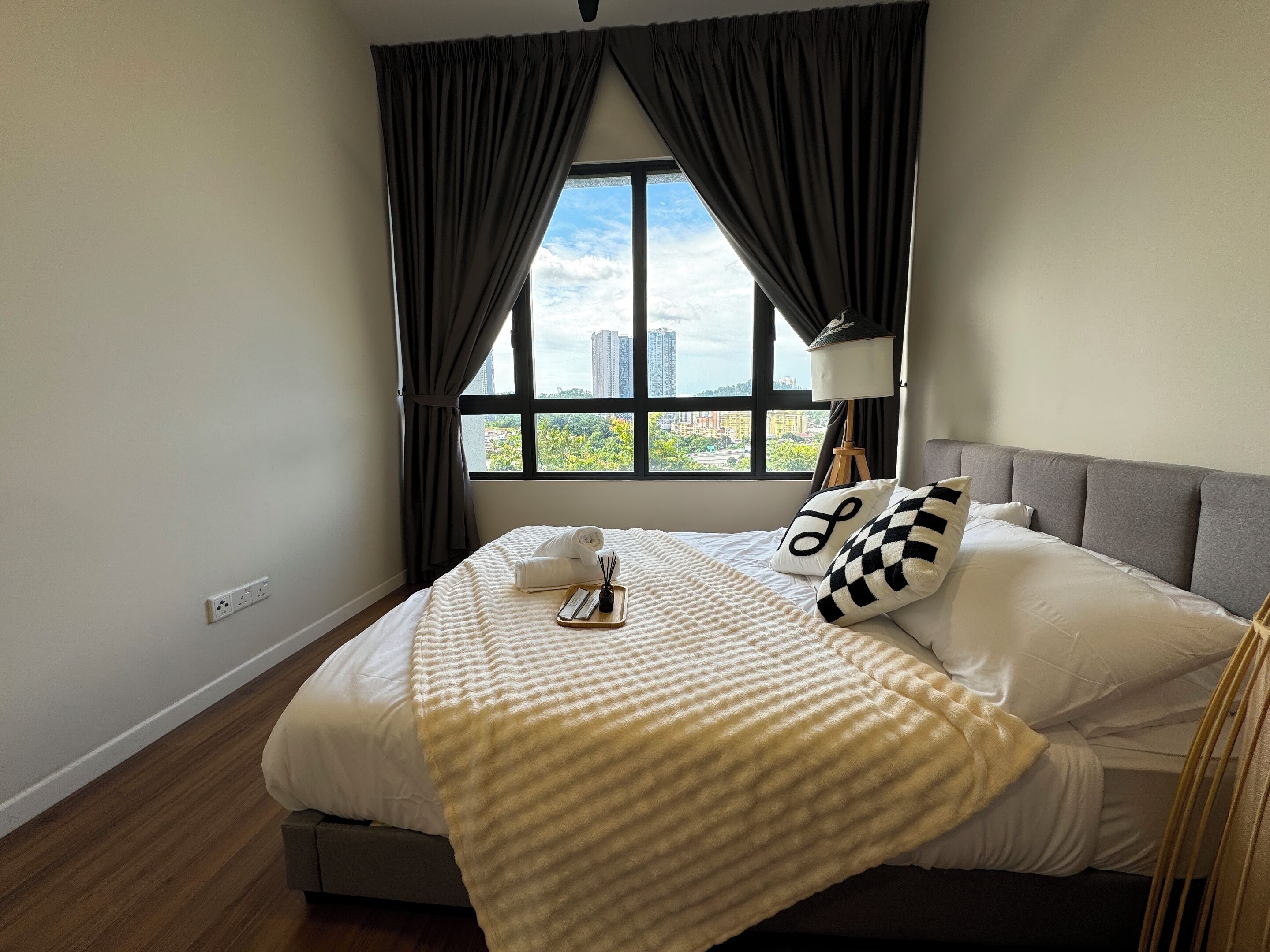 A neatly made bed with a white comforter, a striped blanket, and a pillow with a checkered pattern. There is a bedside table with a lamp and a tray with a remote control. The room has a large window with curtains, and a cityscape is visible outside.