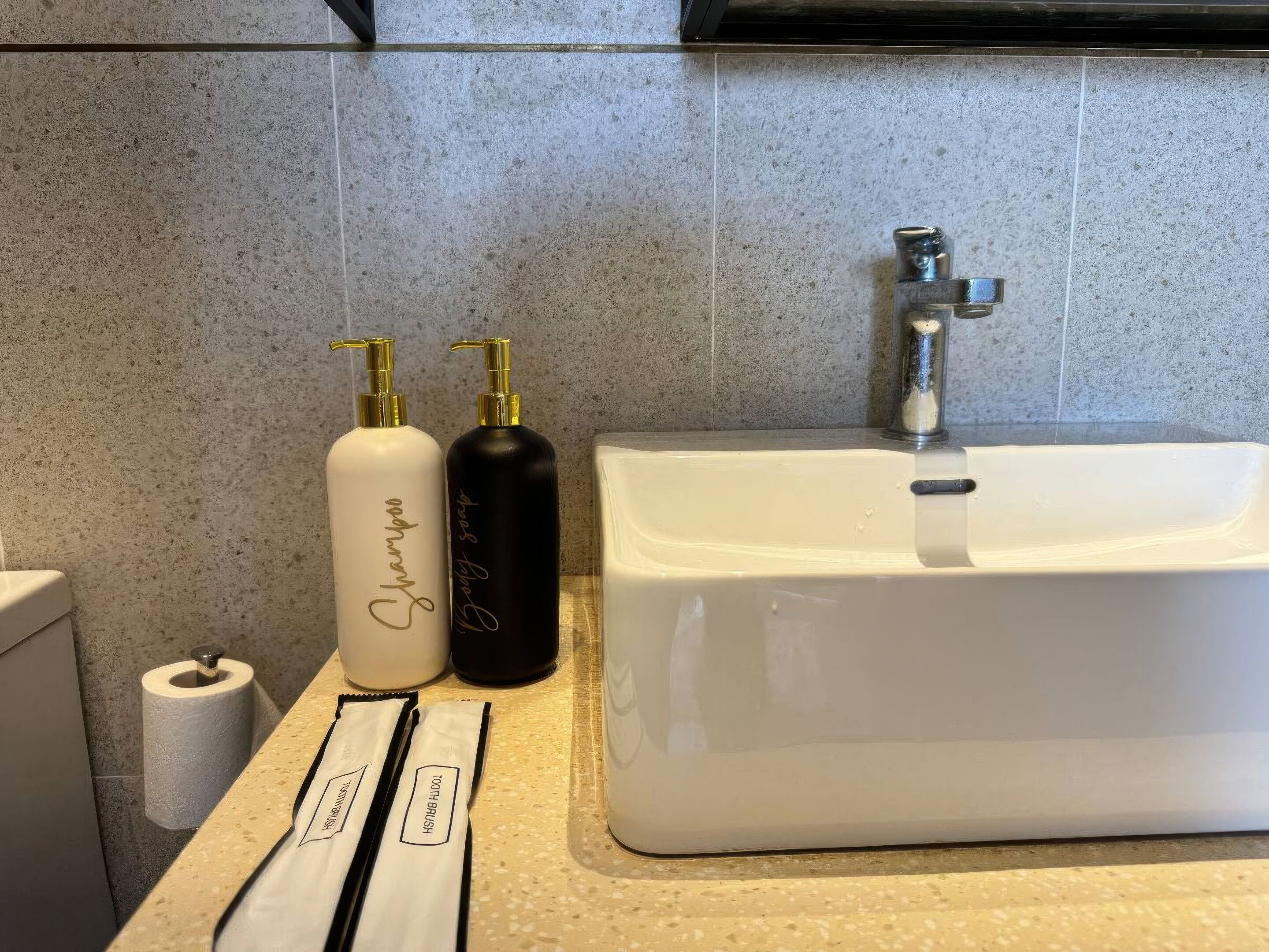 Un bagno con un lavandino bianco, due flaconi di articoli da toilette e un rotolo di carta igienica.