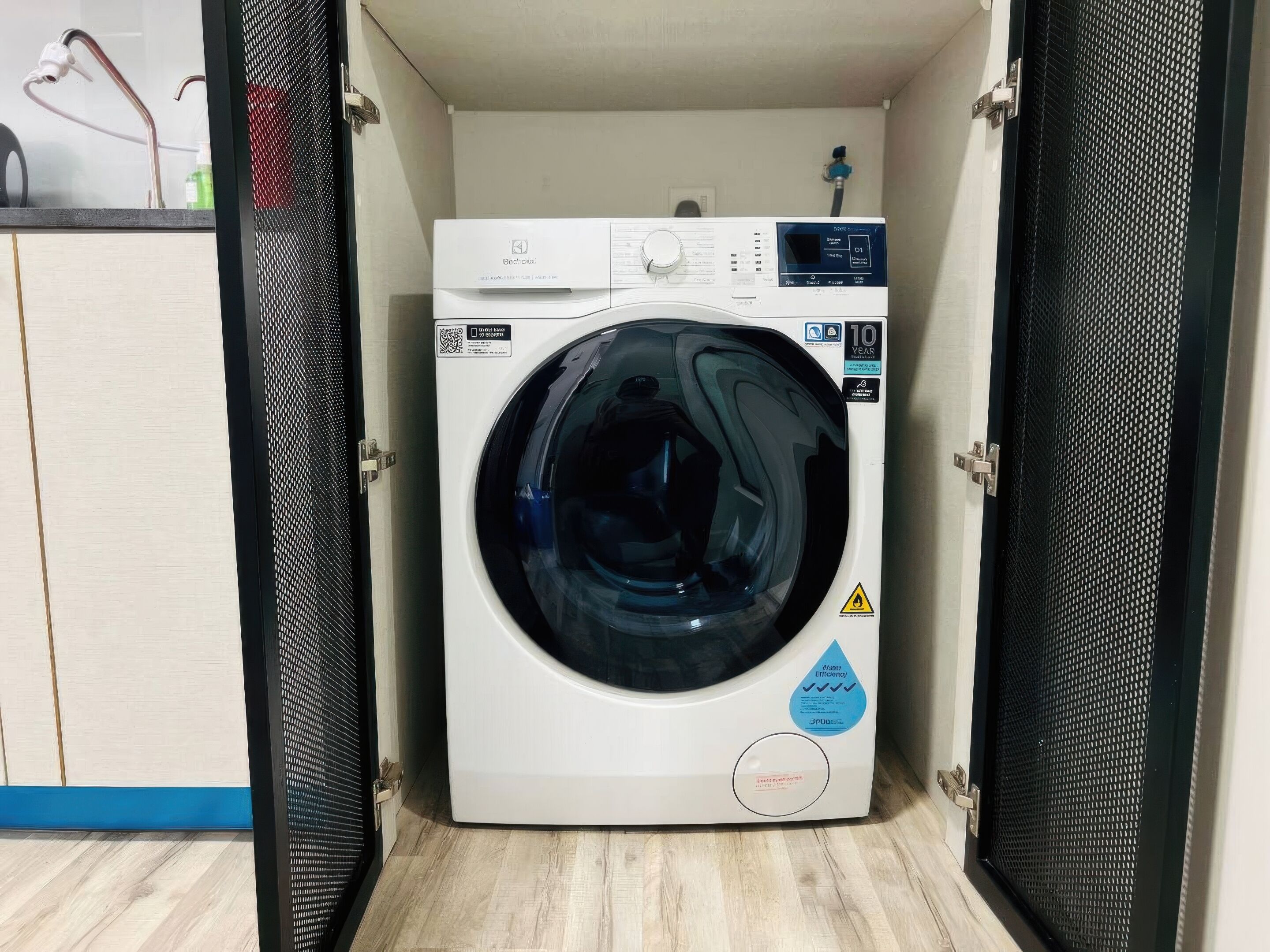 Een compacte, witte wasmachine voorraam met een digitaal scherm, geplaatst in een kleine wasruimte met houten vloer en blauwe afwerking.